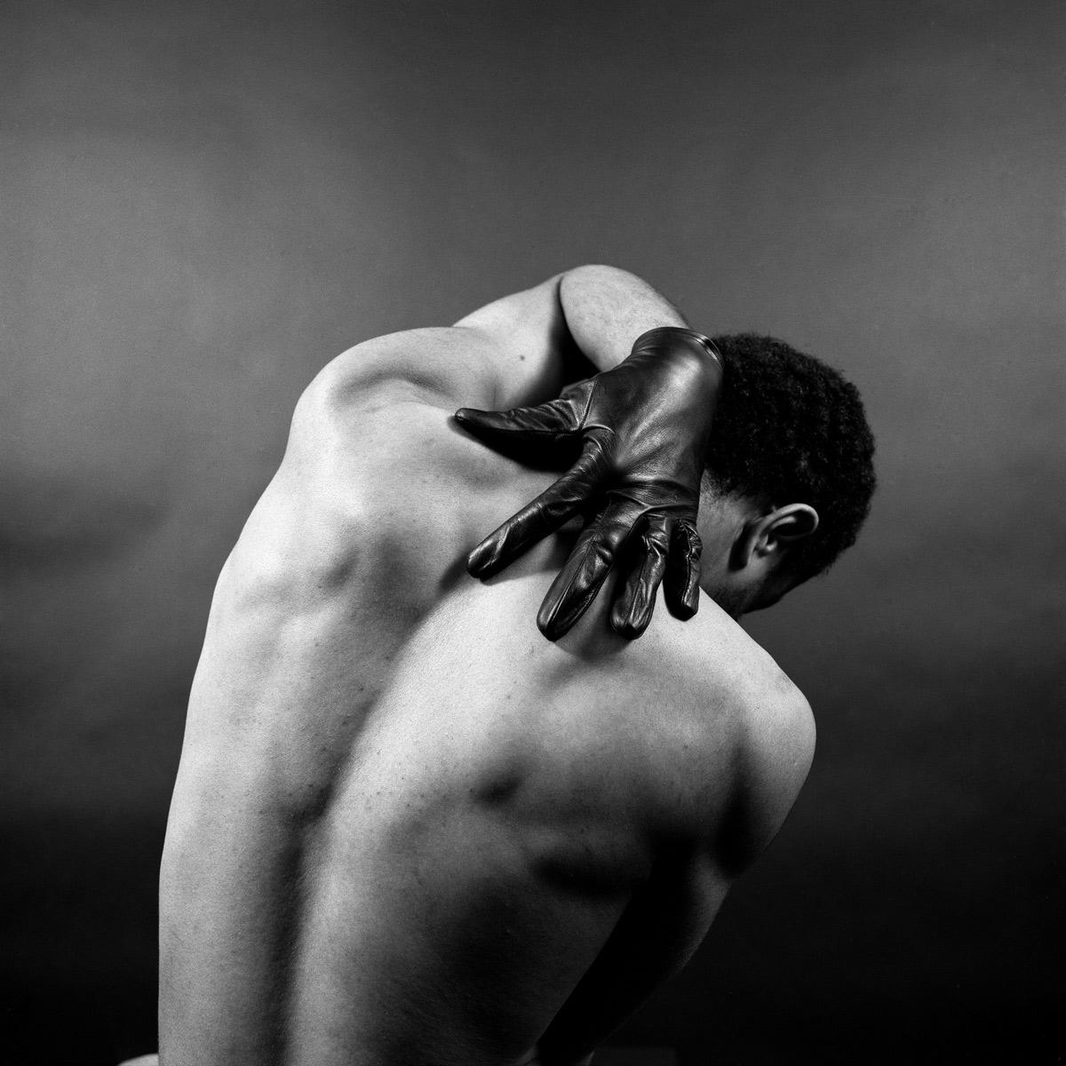 Black and white photograph showing the back of a Black man. One hand wearing a black glove stretches behind him.