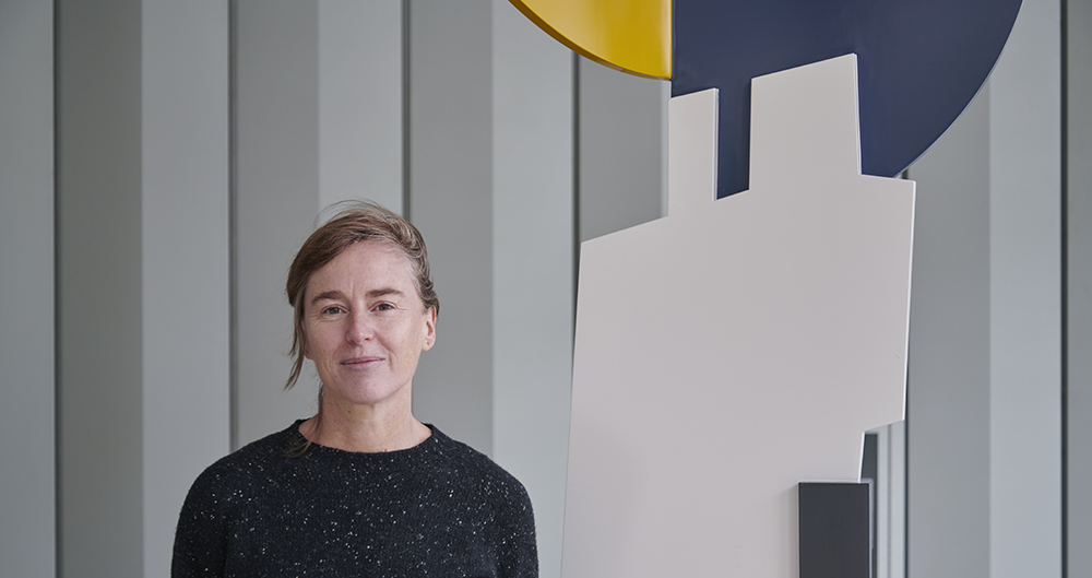 The Artist Fiona Curran standing next to one of her public sculptures. The artist is a white presenting person wearing a black top standing against a grey neutral background and next to a sculpture made up of white, yellow and navy colour blocks.