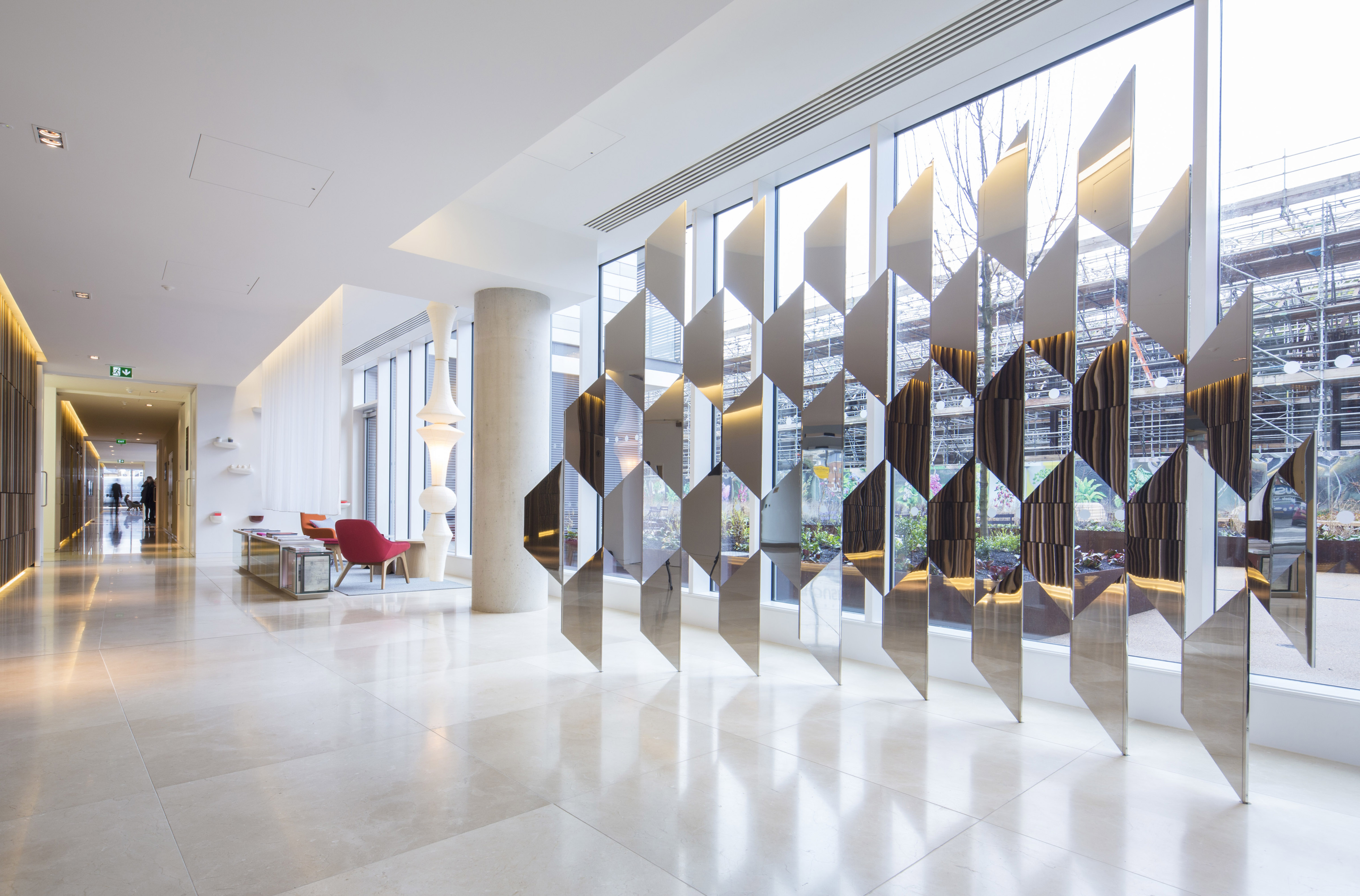 An install shot of an artwork by artist Robert Orchardson. The image depicts two mirror sculptural forms which interlock to create a shifting and repeating hexagonal pattern. The sculpture is located in an office lobby space set against a large glass window.